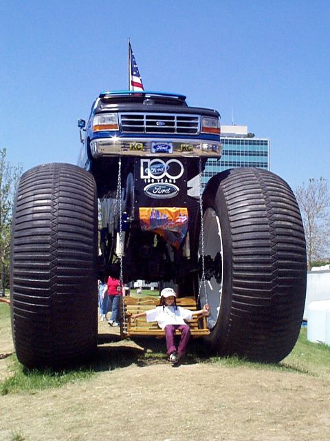 1999 Ford Taurus - A monster truck, likely a Ford, is displayed with oversized tires and a "100 Years Ford" badge, while a child sits on a swing beneath it.