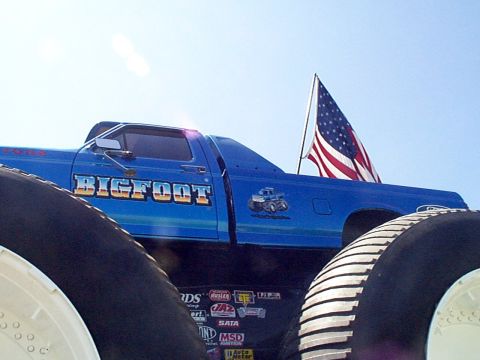 1999 Ford Taurus - A blue Bigfoot monster truck is viewed from a low angle against a clear sky, with an American flag displayed.
