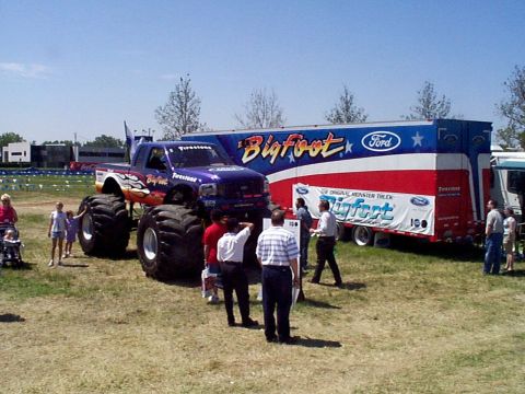 1999 Ford Taurus - Bigfoot monster truck and trailer on display with onlookers.