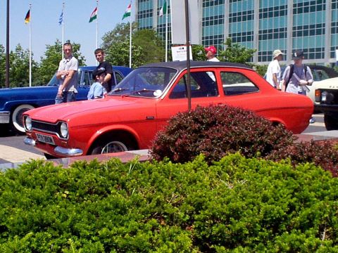1999 Ford Taurus - A red coupe with a black roof is parked in front of a modern building with flags flying.