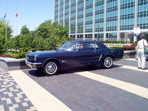 1999 Ford Taurus - A dark blue 1965 Ford Mustang coupe is parked on a brick walkway in front of a modern building.