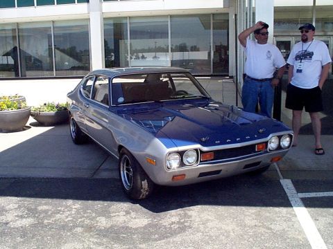 1999 Ford Taurus - A two-tone Ford Capri is parked in front of a modern building, with two onlookers.