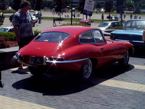 1999 Ford Taurus - A red 1963 Jaguar E-Type coupe is parked on a brick surface, with a man standing nearby and other vehicles in the background.