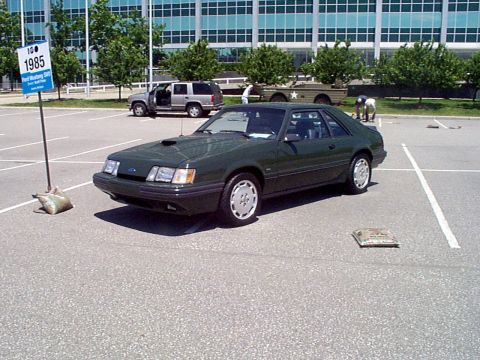 1999 Ford Taurus - A dark green 1985 Ford Mustang coupe is parked in a lot next to a sign indicating its year.