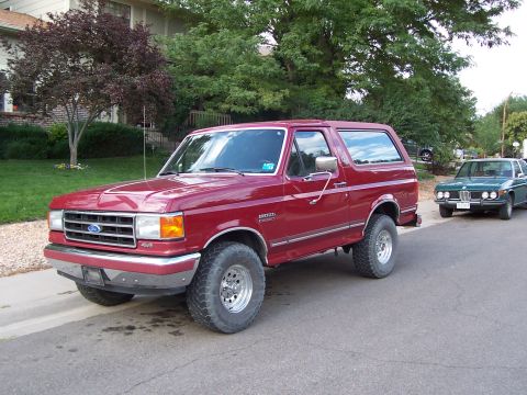 1991 Ford Bronco - A red 1991 Ford Bronco is parked on a residential street next to a green sedan.
