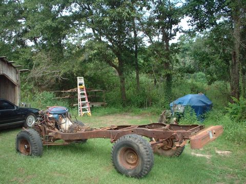 1976 Ford F-150 - A weathered truck chassis sits on grass, showing its engine and a flat front tire, with a blue tarp-covered object in the background.