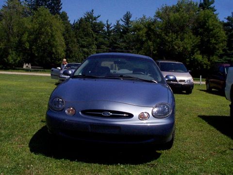 1999 Ford Taurus - Front view of a blue 1999 Ford Taurus parked on grass with other vehicles in the background.
