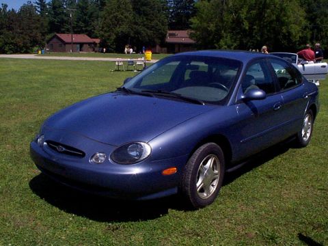 1999 Ford Taurus - A blue 1999 Ford Taurus sedan is parked on a grassy area with trees and buildings in the background.