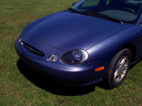 1999 Ford Taurus - The front end of a blue 1999 Ford Taurus is visible, parked on a grassy area.