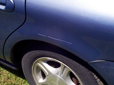 1999 Ford Taurus - Scratches mar the dark blue paint on the rear quarter panel of a car, with a silver wheel visible below.