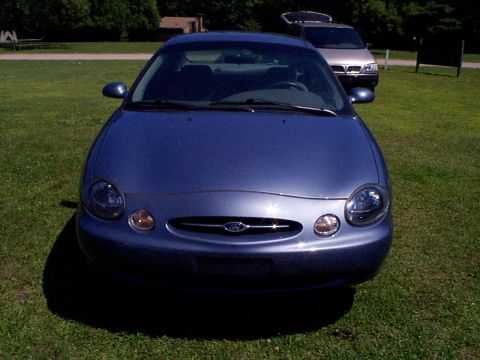 1999 Ford Taurus - A blue 1999 Ford Taurus sedan is parked on a grassy area with another vehicle visible behind it.