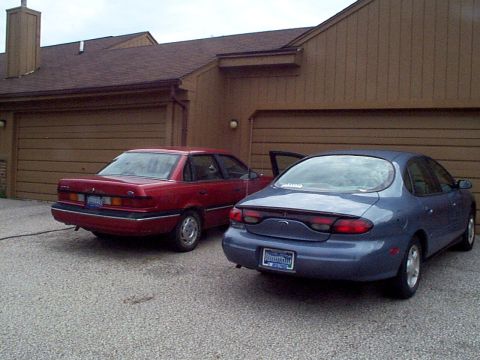 1999 Ford Taurus - Two sedans are parked in a driveway in front of a house with two garage doors.