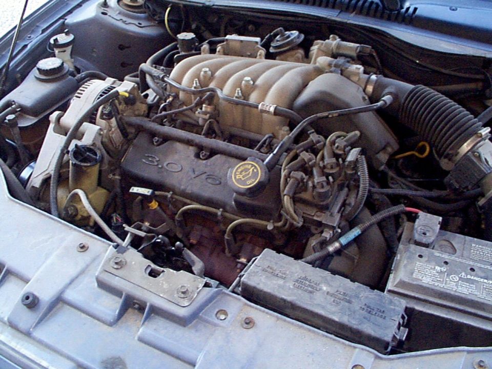 Engine bay of a 1999 Ford Taurus showing the 3.0 V6 engine, battery, and fuse box