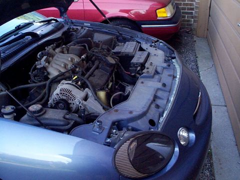1999 Ford Taurus - The engine bay of a 1999 Ford Taurus SE is shown, with a red vehicle parked behind it.