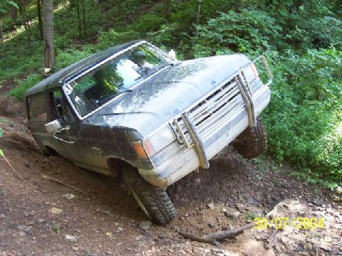 1995 Ford Bronco - A dark blue Ford Bronco is shown off-road, tilted on a muddy slope with trees in the background.