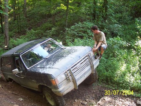 1995 Ford Bronco - A dark-colored SUV is stuck in the mud on a wooded trail, with a man sitting on its hood.