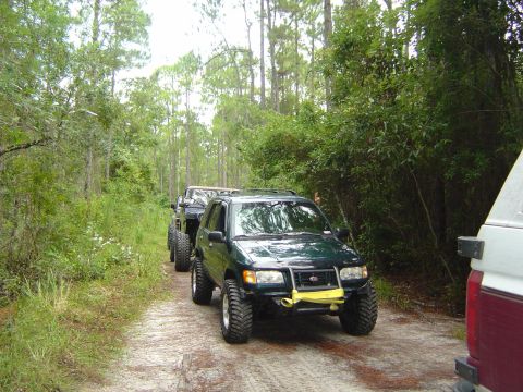 1993 Ford Bronco - Multiple off-road vehicles drive down a dirt road surrounded by trees.