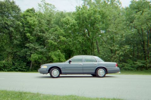 1998 Ford Crown Victoria - A 1998 Ford Crown Victoria is seen from the side, parked on a road with a wooded area behind it.