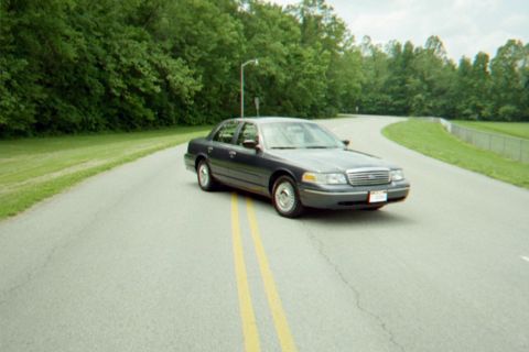 1998 Ford Crown Victoria - Dark colored 1998 Ford Crown Victoria on a paved road surrounded by greenery.