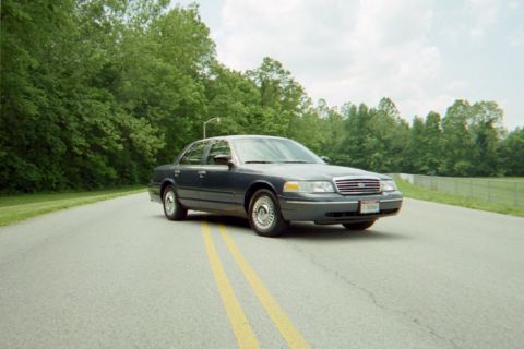 1998 Ford Crown Victoria - A dark-colored 1998 Ford Crown Victoria is shown driving on a paved road surrounded by trees.