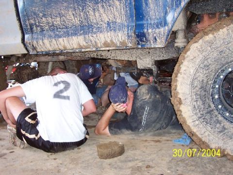 1995 Ford Bronco - People are working on the underside of a muddy vehicle, with a large tire on the right side of the frame.