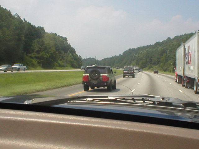 Highway view from inside a vehicle, following a green SUV with rear spare tire; FedEx trailer at right