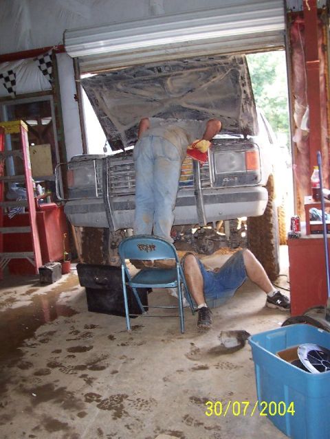 1995 Ford Bronco - Two men are working on a dirty vehicle in a garage, with the hood open and one man lying underneath.