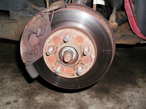 1992 Ford Taurus - A front brake assembly with the wheel off, showing a heavily rusted and grooved five-lug rotor with a visible crack through the outer edge,…
