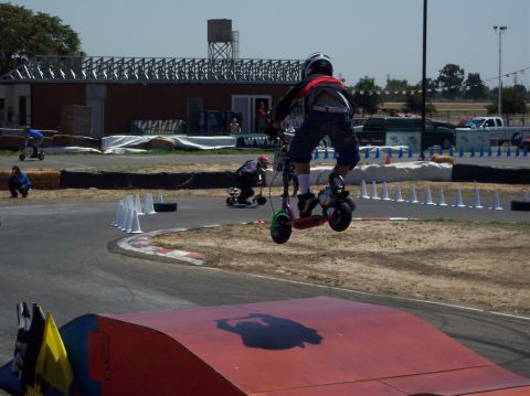 1990 Ford Bronco - Rider on a scooter takes flight over a ramp during a race event. Other participants are visible on the track.