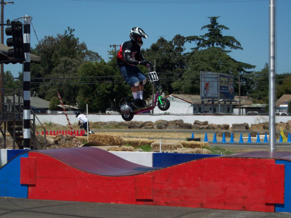 Person on a scooter mid-air over a ramp, wearing a helmet and pads. Number 832 is visible on the scooter