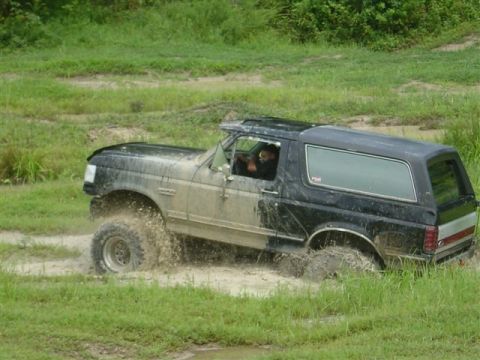 1995 Ford Bronco - Driver-side action shot of a mud-splattered black Ford Bronco blasting through a shallow water hole in a grassy mud pit, throwing spray off…