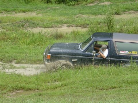 1995 Ford Bronco - Dark blue Ford Bronco with its doors removed runs through a grassy mud pit, front tire kicking up a sheet of muddy water. The driver leans…