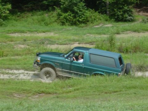 1995 Ford Bronco - Driver-side profile of a teal-green Ford Bronco running through a grassy mud pit with its doors removed, the driver and a passenger visible…