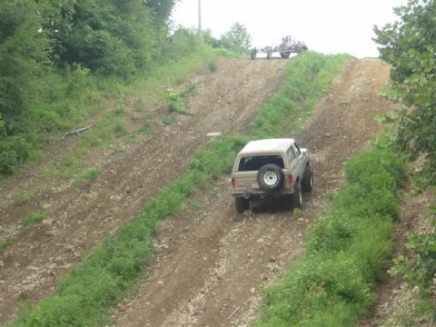 1995 Ford Bronco - Rear three-quarter view of a tan full-size Ford Bronco partway up a steep, rutted dirt hillclimb, with a spare tire mounted on the rear bod…