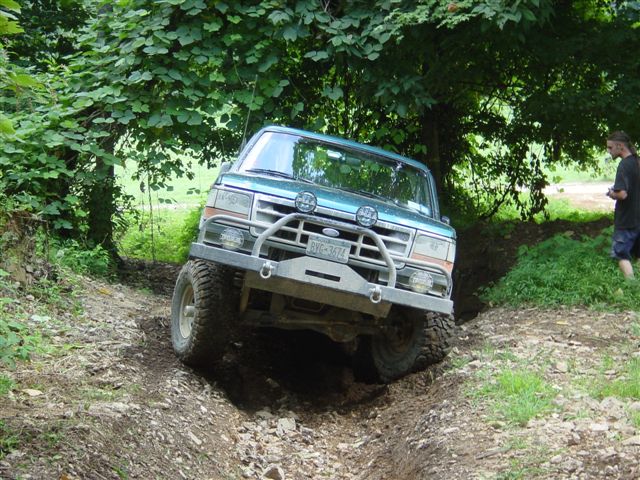 Lifted teal Ford Bronco with tube bumper and winch flexing in a muddy off-road rut