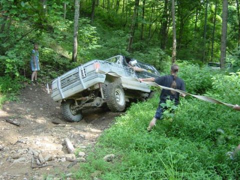 1995 Ford Bronco - A white/silver SUV is stuck at a steep angle on a rocky off-road trail in the woods, with its front passenger wheel dropped into a rut, whi…