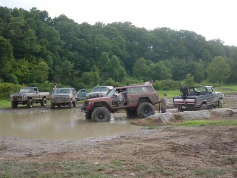 1995 Ford Bronco - Off-road event at a mud pit ringed by wooded hills: a mud-covered lifted SUV with its doors removed climbs out of the water in the foregrou…
