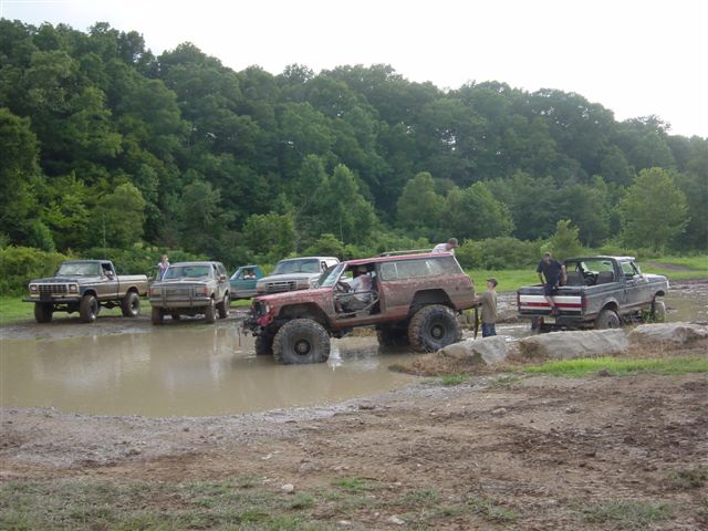Mud-covered lifted SUV exiting a mud pit at an off-road event with trucks waiting behind
