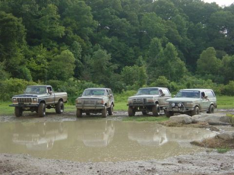 1995 Ford Bronco - Four lifted Ford trucks parked side by side in a muddy off-road lot, including two older Ford pickups and two Bronco SUVs, all facing the c…