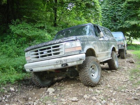 1995 Ford Bronco - A mud-splattered white and dark-colored Ford Bronco full-size SUV crawling over a rocky off-road trail, shown from the front three-quarter…