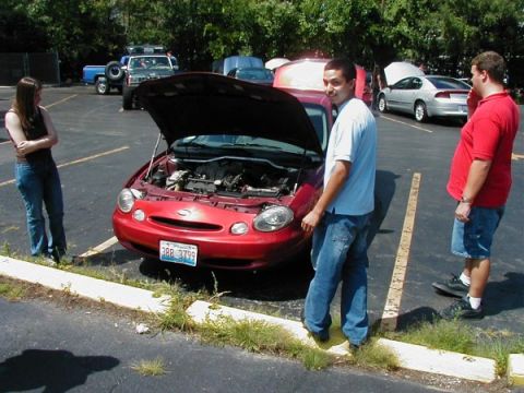 1997 Ford Taurus - People gathered around a red 1997 Ford Taurus with its hood open in a parking lot.