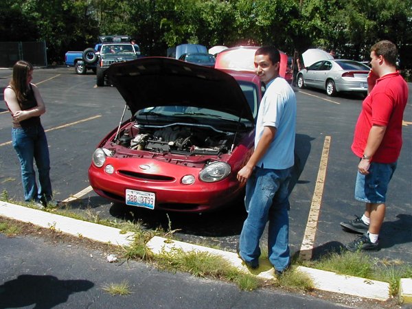 Red 1997 Ford Taurus with hood open, license plate 388 3799. Two men and a woman stand nearby in a parking lot
