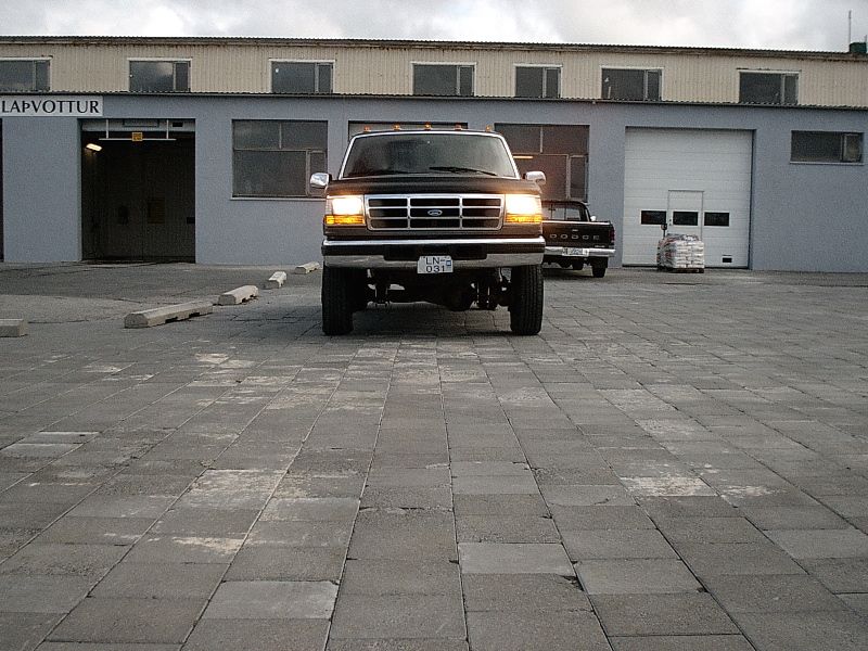 Dark Ford truck seen from front with lights on, parked outside a gray industrial building.
