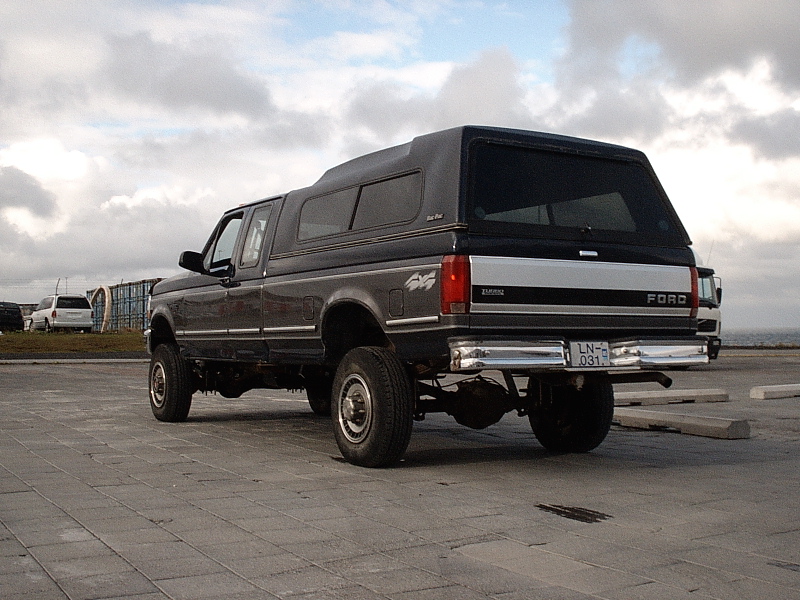 Rear 3/4 view of a lifted dark Ford 4x4 pickup with camper shell on a paved lot