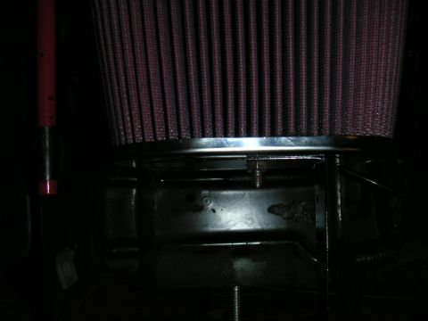 1994 Ford Bronco - Dim close-up of the end cap of a pink/red pleated cone air filter with a chrome ring, mounted above a black sheet-metal bracket in an engin…