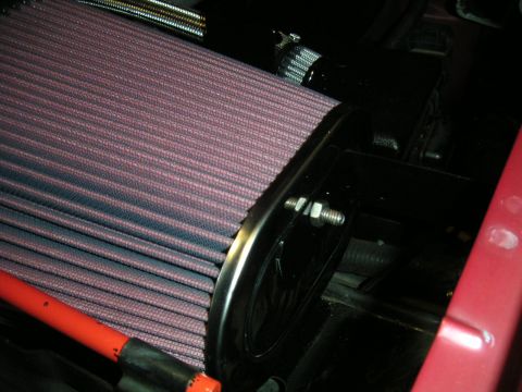1994 Ford Bronco - Tight close-up of a red pleated cotton-gauze cone air filter with a black rubber base flange, mounted inside an engine bay. A red spark plu…