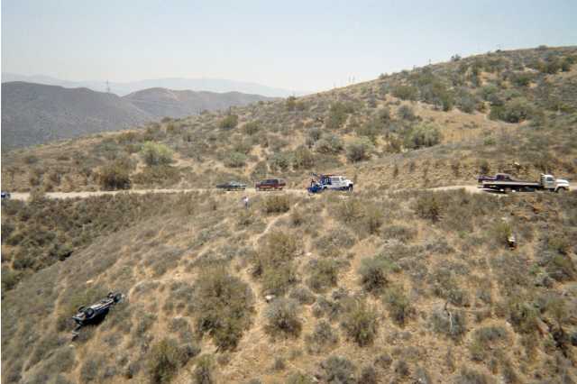 Aerial view of an overturned dark vehicle on a brushy hillside below a dirt road