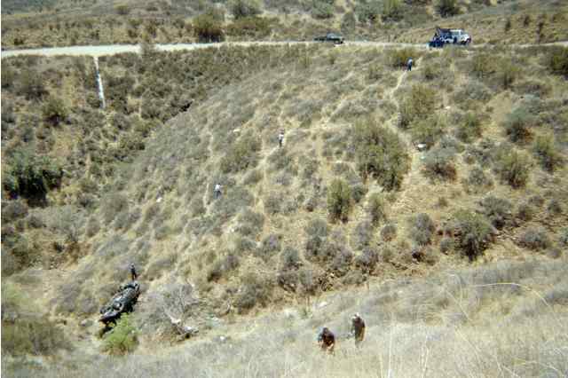 People walking down a brushy hillside toward a wrecked vehicle below a dirt road