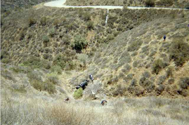 Distant overturned vehicle in a brushy ravine with people on the slope
