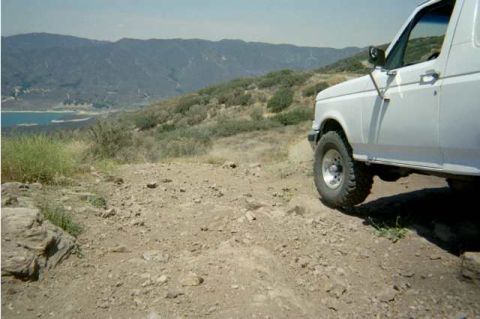 1990 Ford Bronco - front fender and lifted wheel of a white truck on a rocky ridge, overlooking a reservoir and forested mountains below.